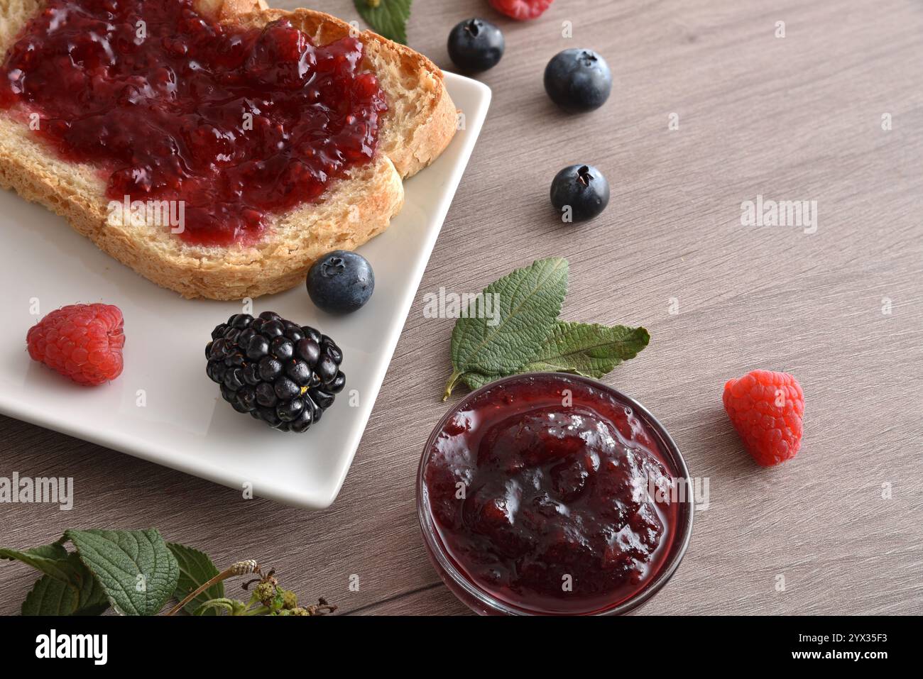 Dettaglio del toast con marmellata di frutta della foresta su un piatto bianco su una panchina di legno con vasetti e frutti di bosco. Vista dall'alto in alto. Foto Stock