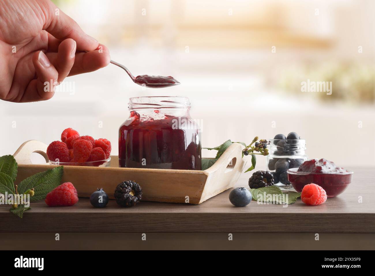 Cucchiaio ripieno a mano con marmellata di frutta di bosco da un vaso pieno su una panchina di legno con frutti di bosco e cucina sullo sfondo. Vista frontale. Foto Stock