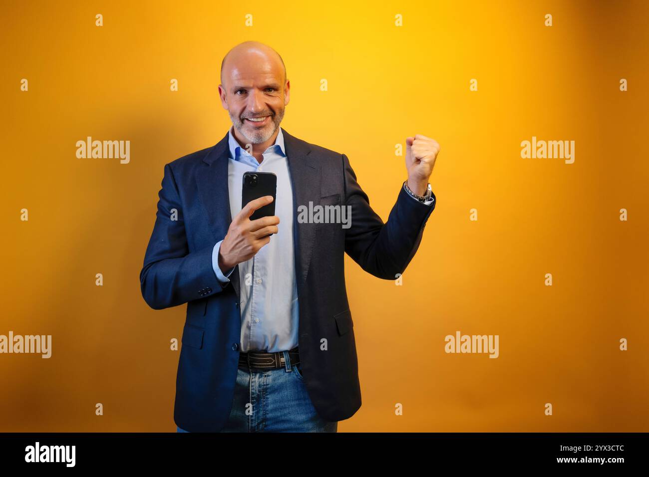 Un uomo con una tuta davanti a uno sfondo giallo. Tiene un telefono nero nella mano destra e alza la mano sinistra in aria. Sorride e guarda Foto Stock