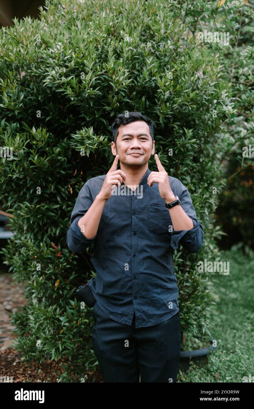 Un uomo del sud-est asiatico che indica giocosamente le sue carine guance, esprimendo gioia e fascino. Perfetto per lo stile di vita, i ritratti o i progetti allegri Foto Stock