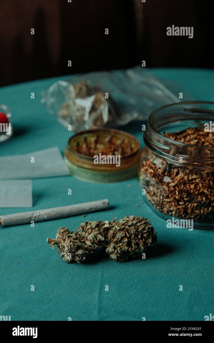 un bocciolo di cannabis su un tavolo accanto a una spliffa, un vaso di vetro con del tabacco arrotolato, alcuni fogli di carta arrotolata, un macinacaffè per erbe usato e un sacchetto di plastica Foto Stock