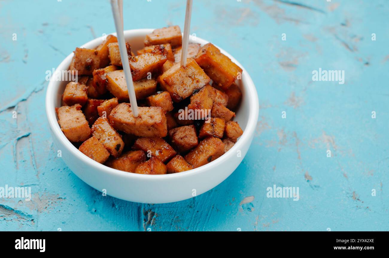 primo piano di una ciotola di ceramica bianca con alcuni cubetti di tofu marinati, servita come spuntino con alcuni spiedini di legno, su una superficie a trama blu Foto Stock