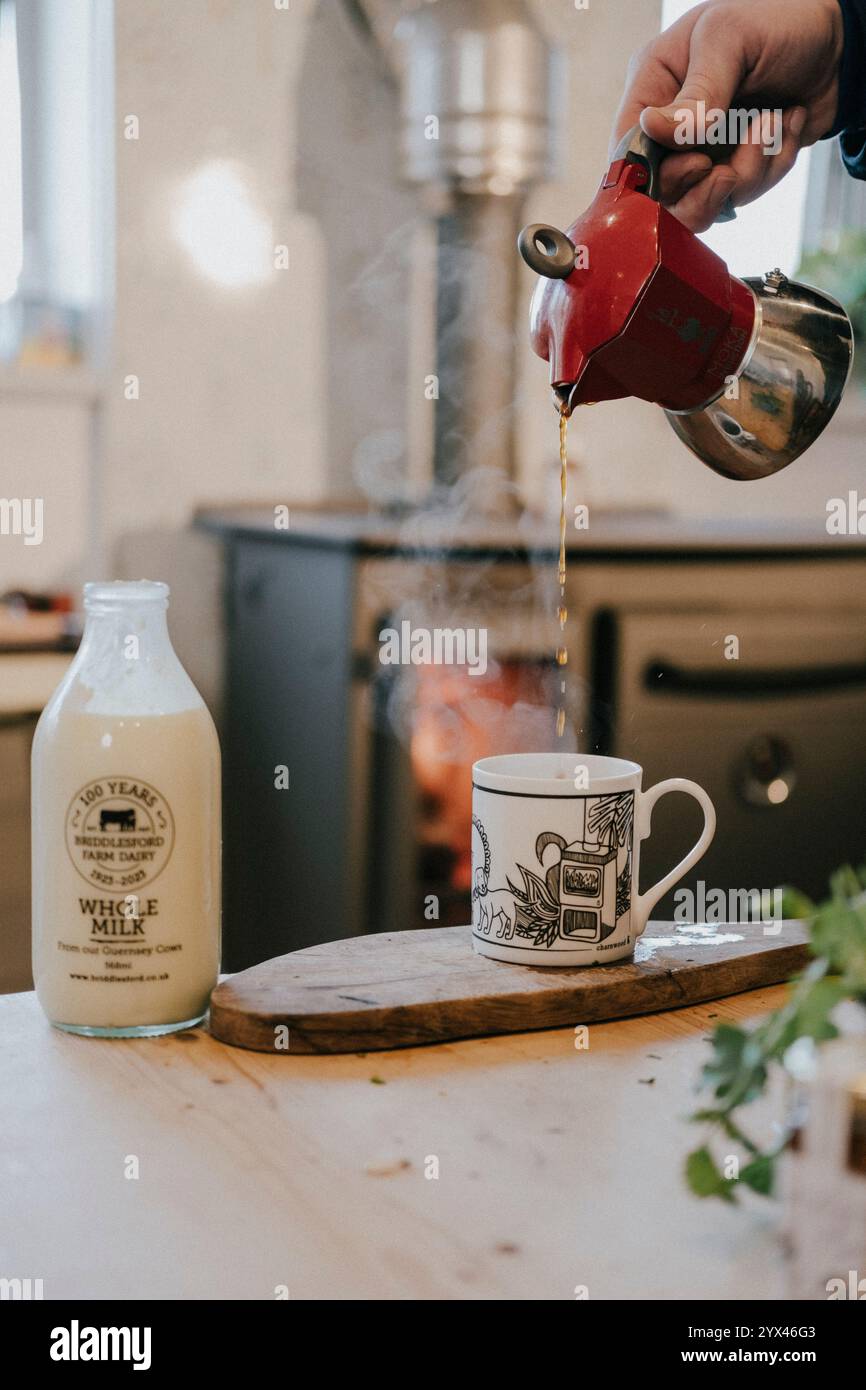 Una persona sta versando il caffè in una tazza da una teiera rossa. La teiera si trova su un vassoio di legno accanto a una bottiglia di vetro. La scena è accogliente e invitante, con t Foto Stock
