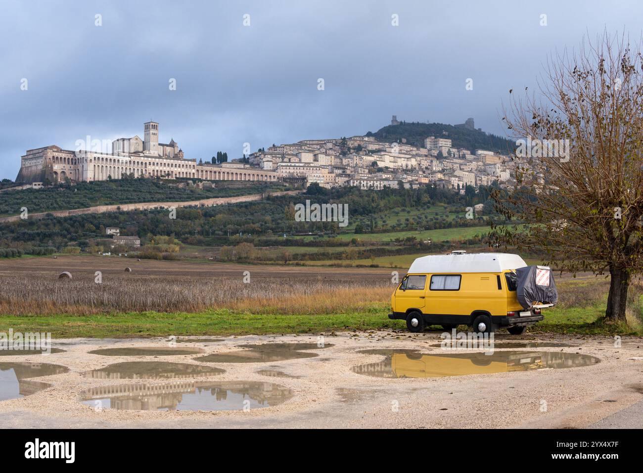 Camper d'epoca giallo parcheggiato di fronte alla città collinare di Assisi in una giornata nuvolosa in Umbria, Italia. Foto Stock