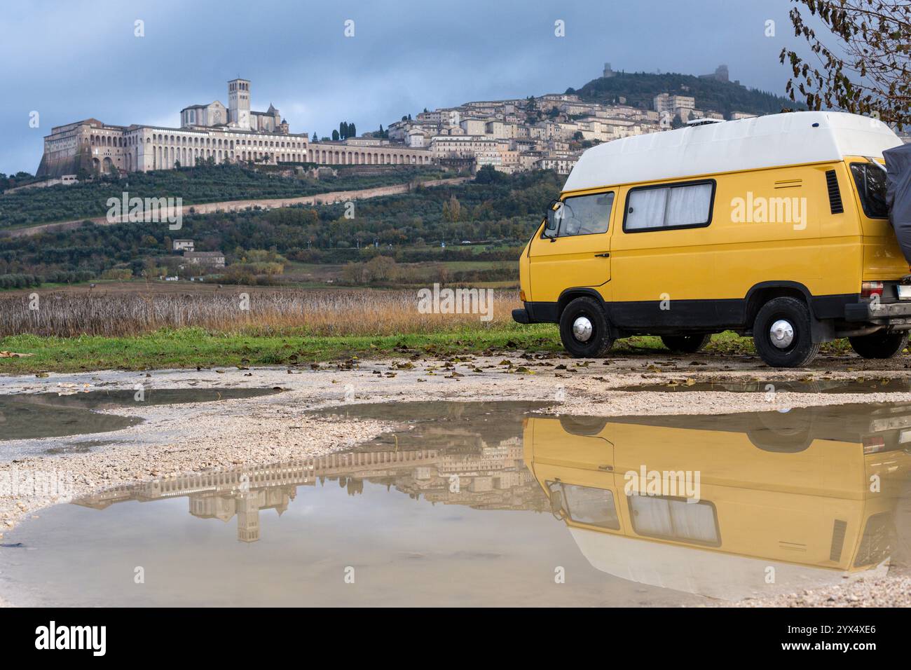 Camper d'epoca giallo parcheggiato di fronte alla città collinare di Assisi in una giornata nuvolosa in Umbria, Italia. Foto Stock