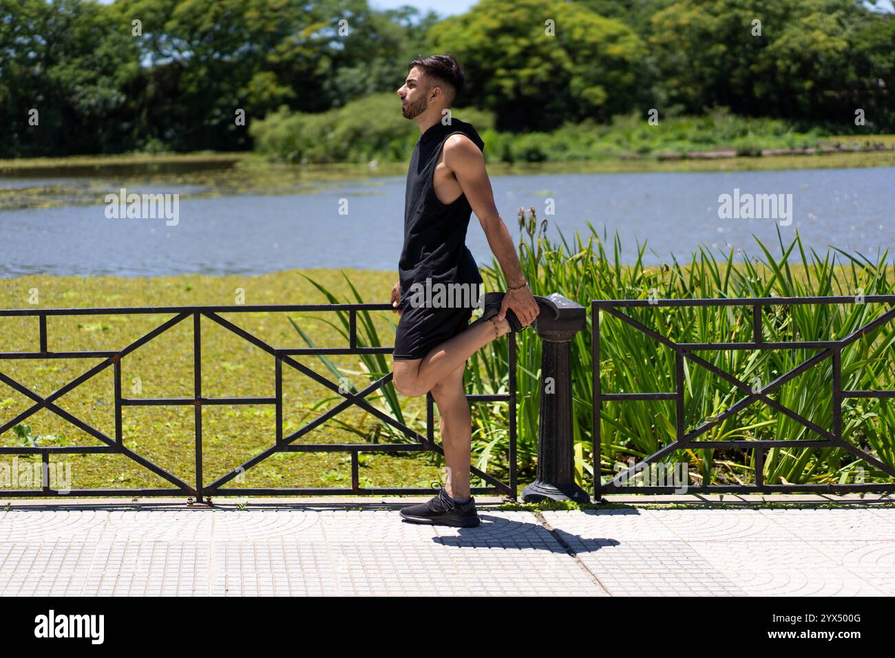 Giovane uomo in forma che allunga il suo quadricipite in riva al lago dopo un allenamento nel parco cittadino. Foto Stock