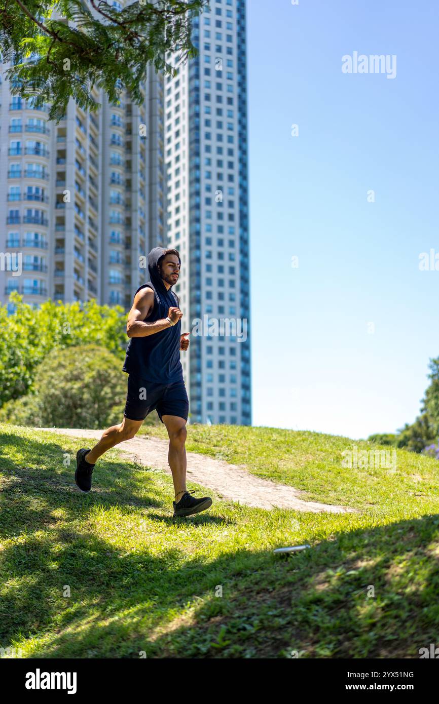Giovane atletico che affronta alti e bassi mentre fa jogging in un parco urbano con edifici contemporanei. Foto Stock