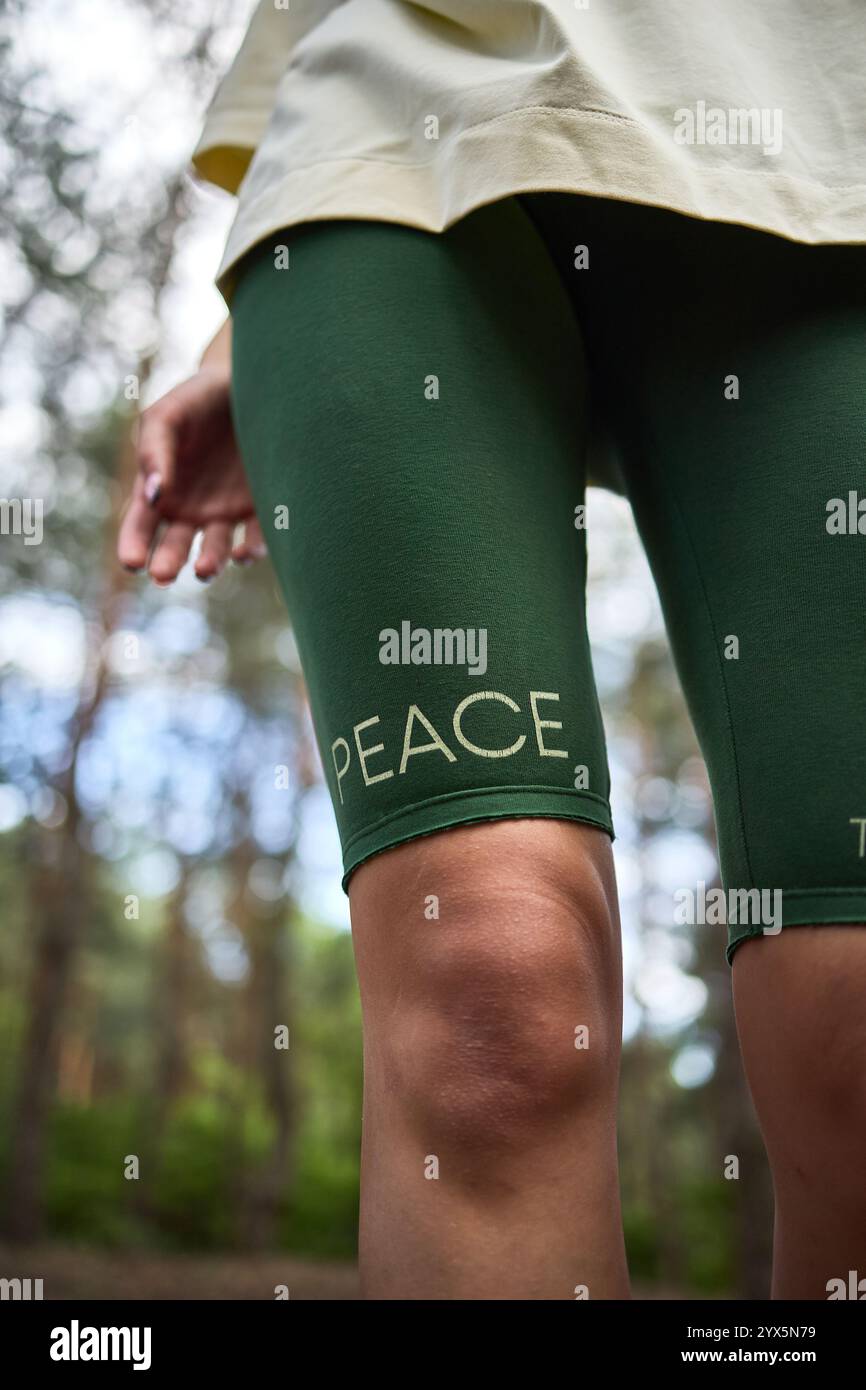 Pantaloncini da ciclismo verdi con un cartello di pace sulle gambe di una donna mentre si trova nella foresta, catturando in primo piano il concetto di stile di vita attivo all'aperto Foto Stock