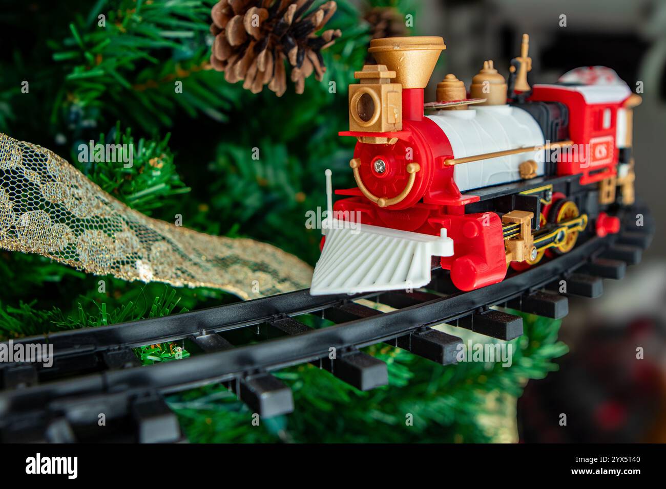 Questa vivace scena natalizia presenta un classico treno giocattolo rosso con accenti dorati che viaggia lungo un percorso tortuoso sotto un lussureggiante albero di Natale verde. Foto Stock
