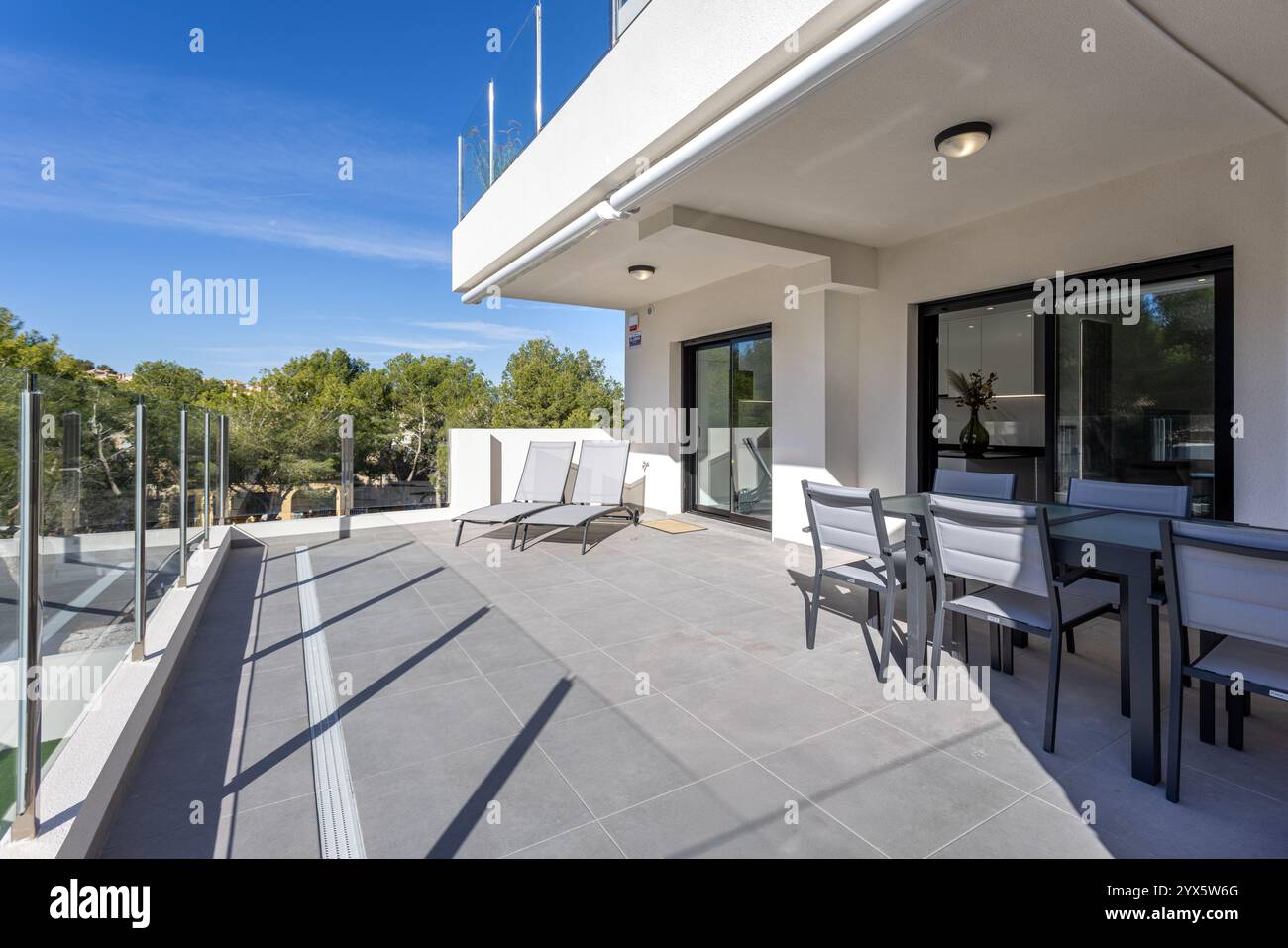 Spaziosa terrazza con eleganti balaustre di vetro, sedie a sdraio e un set da pranzo all'aperto, circondata da serene vedute e luce naturale. Foto Stock