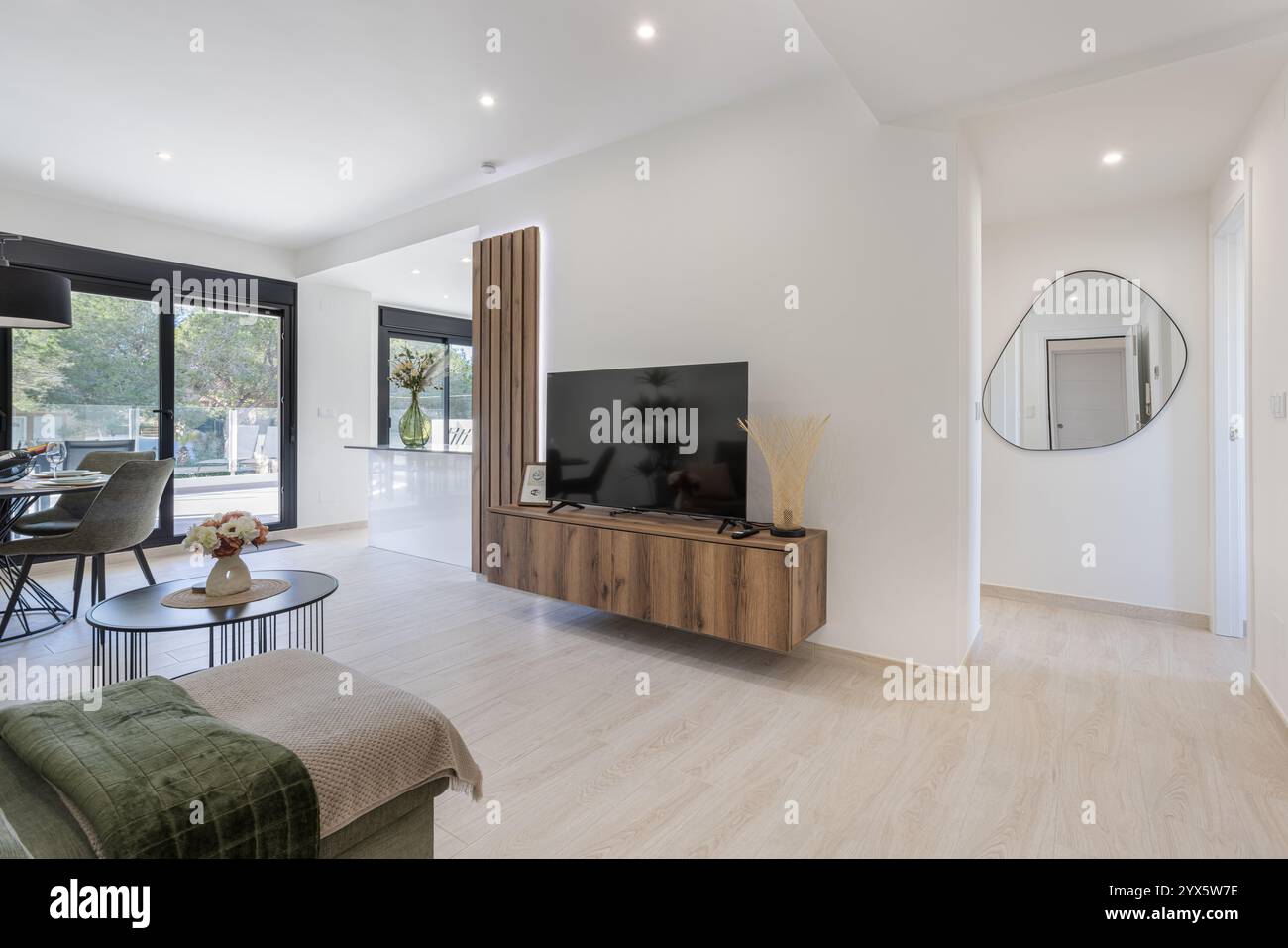 Un soggiorno luminoso e arioso caratterizzato da un'elegante unità TV in legno, zona pranzo, grandi porte in vetro e decor minimalista. Foto Stock