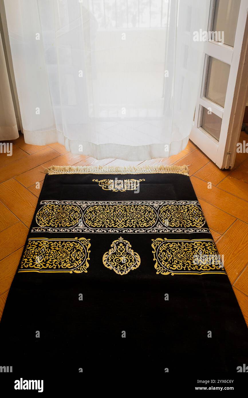Tappeto di preghiera nero islamico con decorazioni dorate, tappeto sul pavimento di fronte ad una finestra con tende bianche, atmosfera spirituale, vibrazioni Ramadan, sfondo Foto Stock