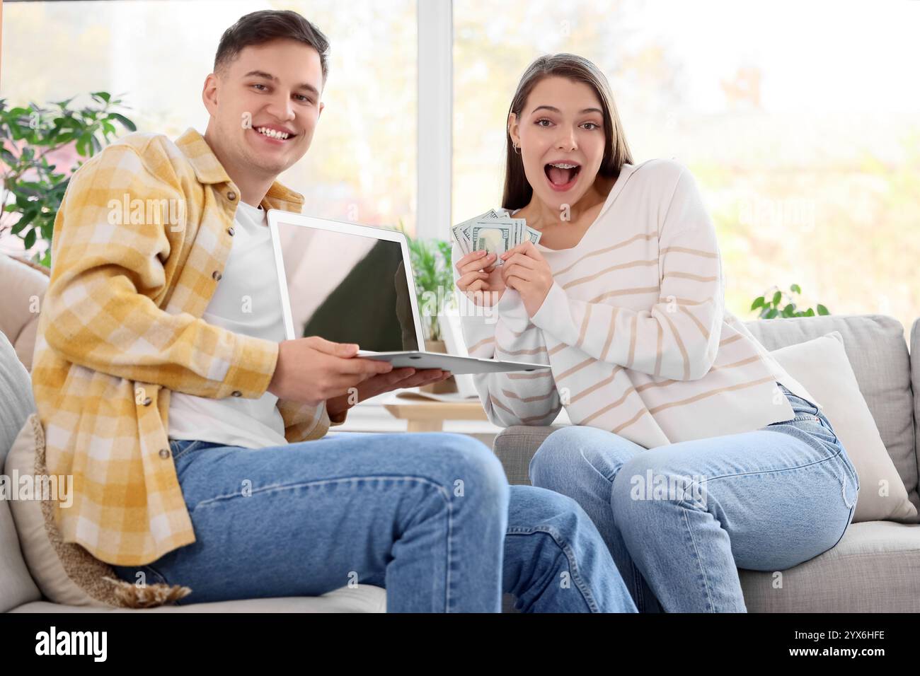 Una giovane coppia felice con soldi e un computer portatile vuoto seduto sul divano di casa Foto Stock