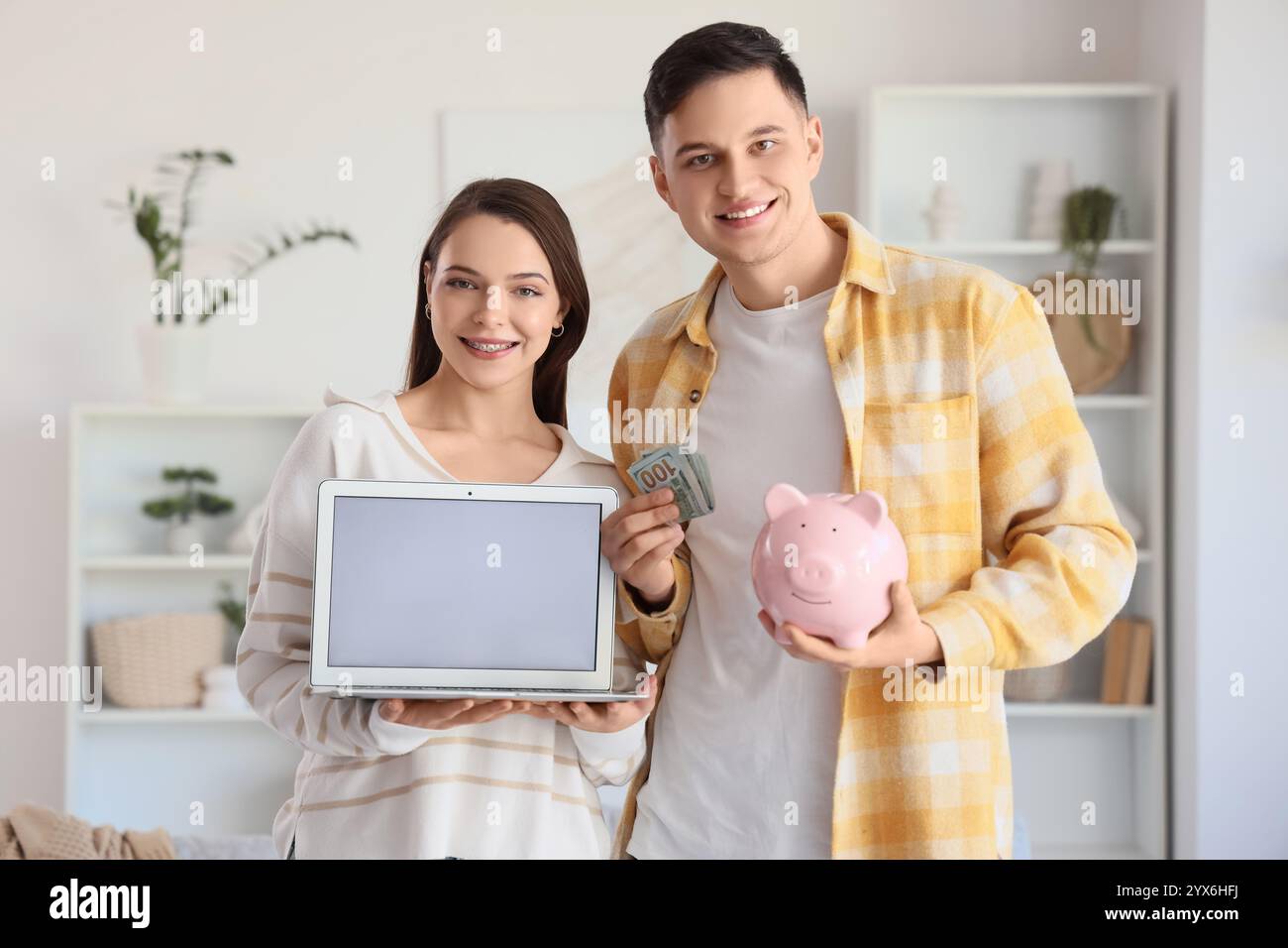 Giovane coppia con soldi, salvadanaio e laptop vuoto a casa Foto Stock