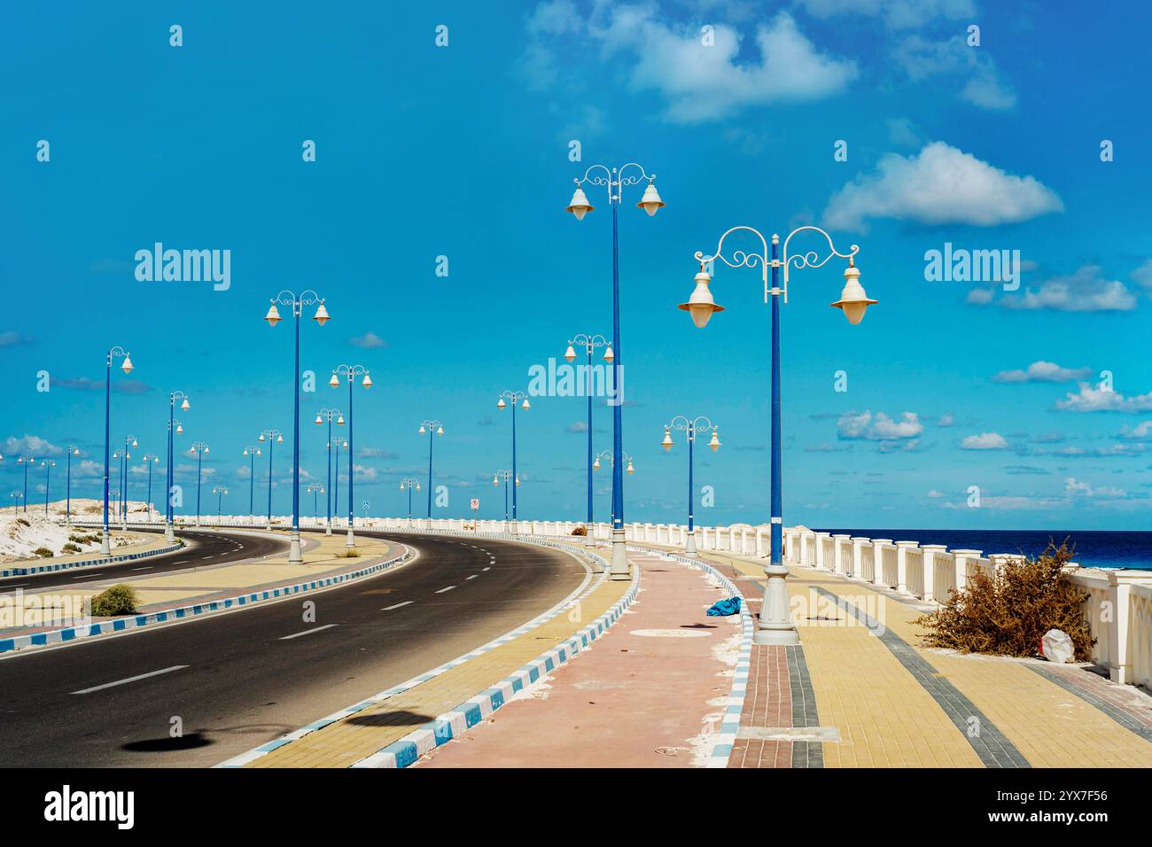 Una pittoresca passeggiata costiera a Mersa Matruh, Egitto, caratterizzata da eleganti lampioni blu, una strada curvilinea e cieli vibranti sul Mediterraneo Foto Stock