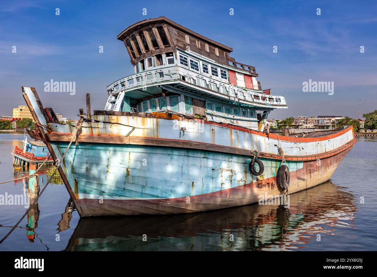 Barca da pesca in legno abbandonata ormeggiata sulla riva del fiume Tha Chin a Bangkok, Thailandia. Fori nel foro, legno rotto, usurato. Cielo blu e nuvole Foto Stock