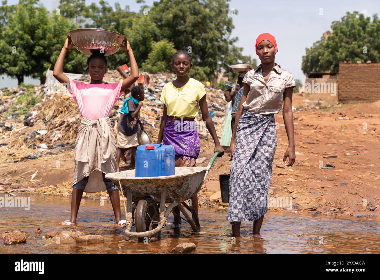 Un gruppo di giovani ragazze del villaggio africano impegnato a raccogliere acqua potabile con una carriola, taniche d'acqua, secchi e ciotole Foto Stock