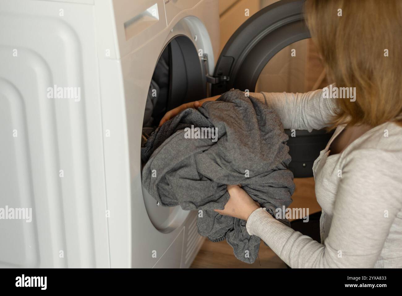 Una donna con un comodo maglione sta mettendo una grande pila di biancheria grigia in una lavatrice a caricamento frontale in una lavanderia ben illuminata. Foto Stock