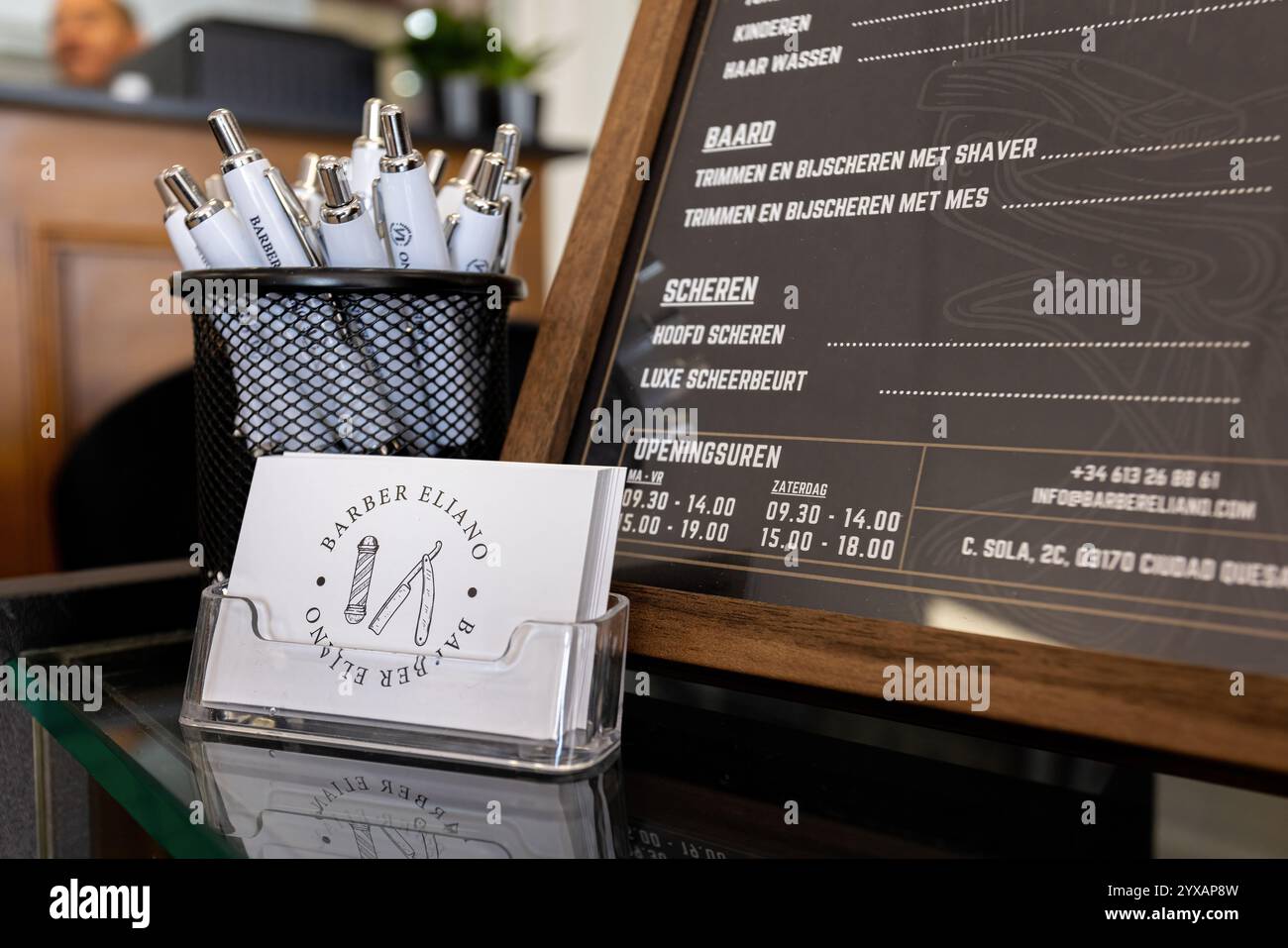 Primo piano di un banco del barbiere con biglietti da visita firmati, penne in un portapacchi e un elegante menu di servizio con orari di apertura. Foto Stock