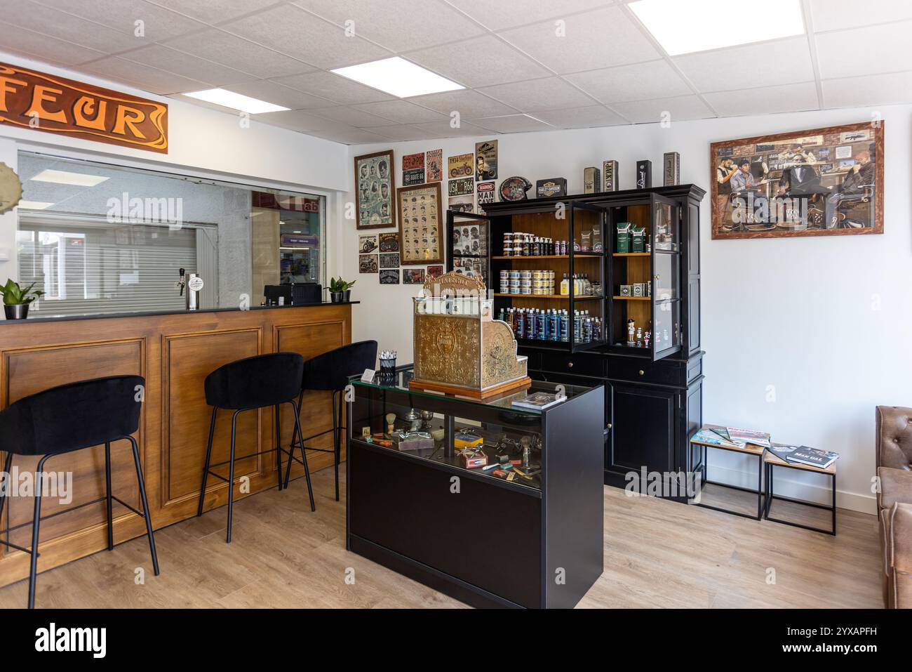 La sala del barbiere presenta un bancone in legno, cassa vintage, scaffali di prodotti, pareti d'arte retrò e comodi posti a sedere. Foto Stock