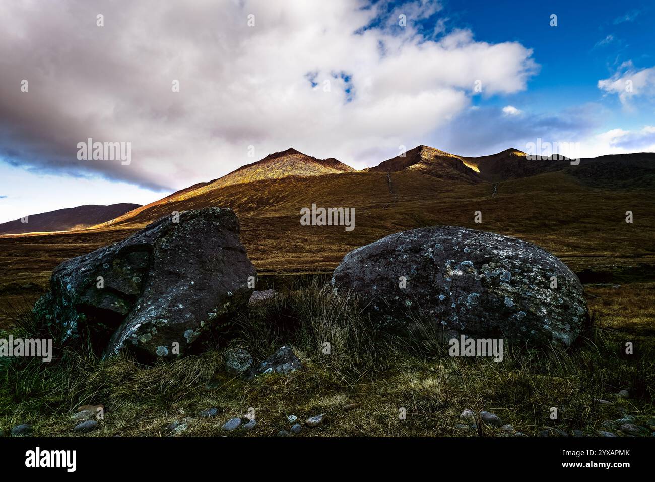 La catena montuosa domina il paesaggio. Due massi poggiano in primo piano. Il terreno è principalmente erba e vegetazione bassa. Foto Stock