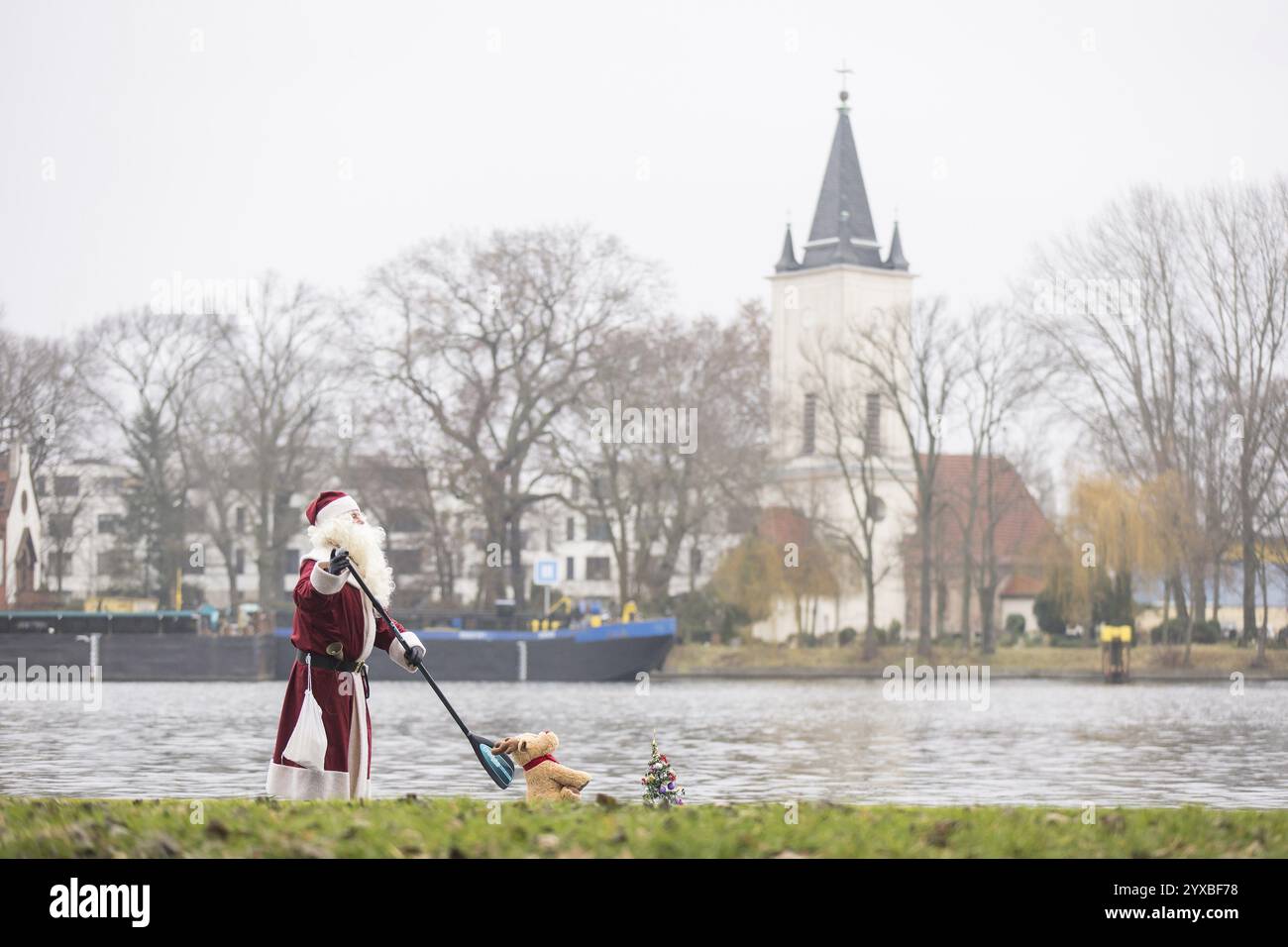 Un appassionato di sport acquatici vestito da Babbo Natale cavalca sup sulla Sprea di fronte alla chiesa del villaggio di Stralau a Berlino il 14 dicembre 2024 Foto Stock