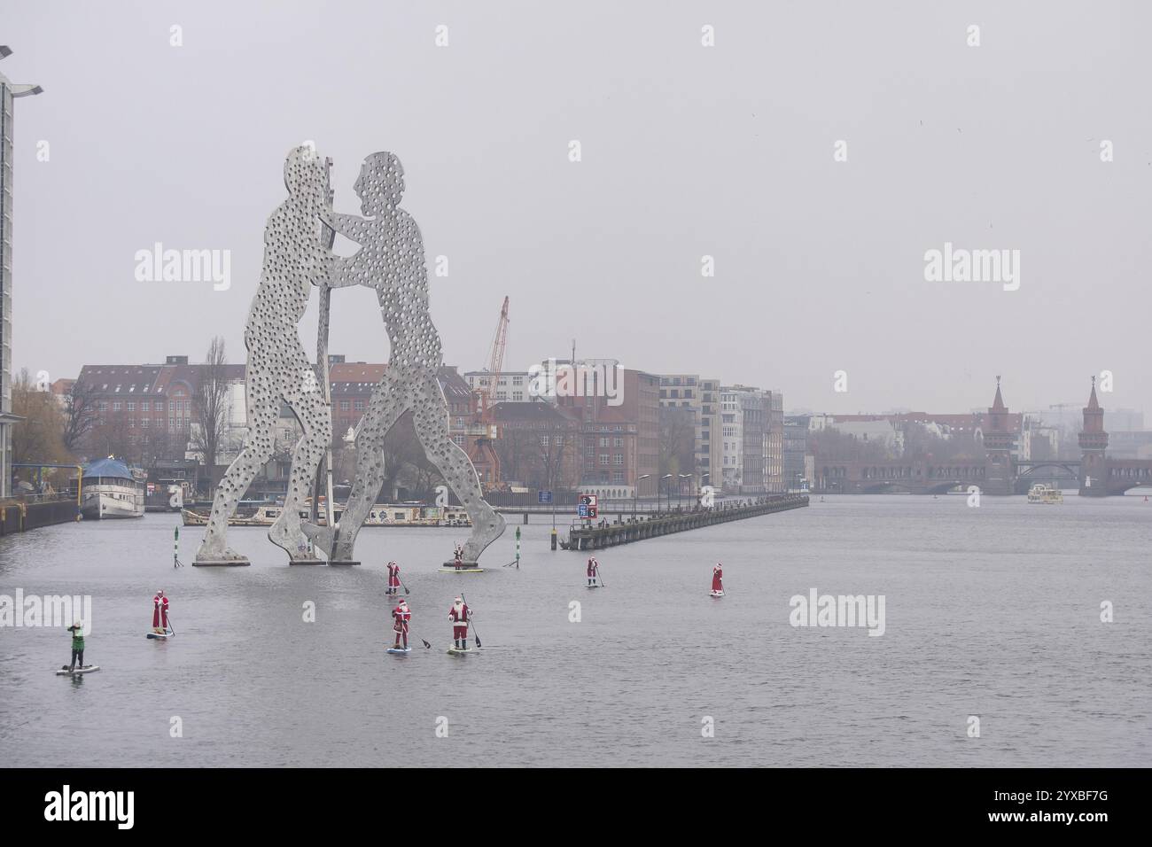 Gli appassionati di sport acquatici, vestiti da Santas, cavalcano SUP sulla Sprea di fronte al Molecule Man e al ponte Oberbaum a Berlino il 14 dicembre 2024 Foto Stock