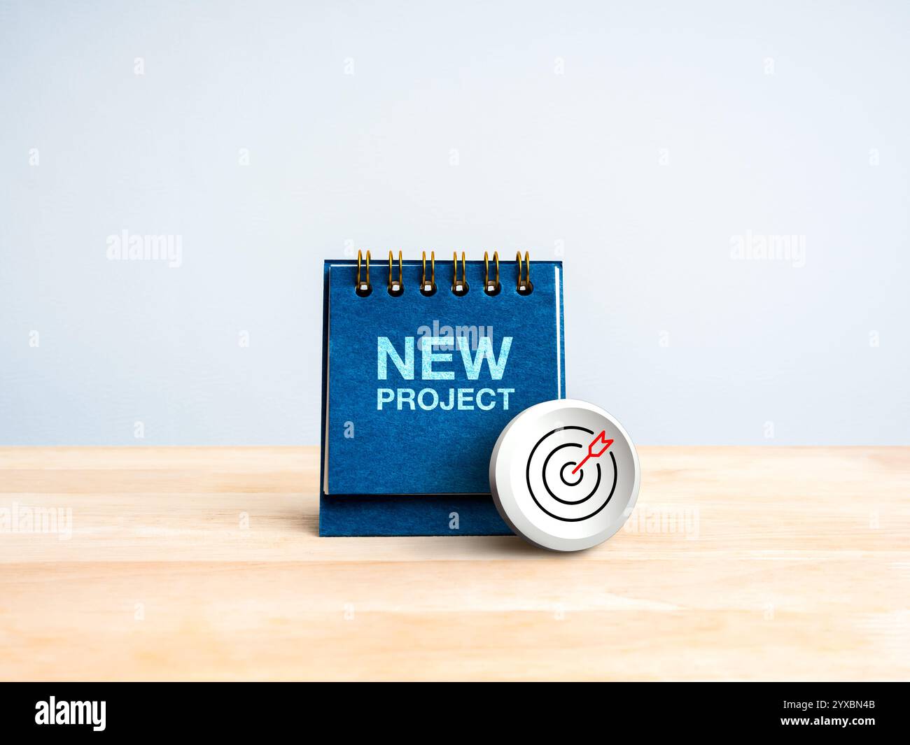 Icona a forma di freccia di destinazione moderna sulla copertina blu del calendario da scrivania con il testo "NEW PROJECT" su tavolo in legno, sfondo bianco. Obiettivo aziendale, obiettivo di successo sc Foto Stock
