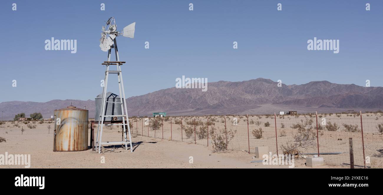 Una pompa d'acqua rustica per mulini a vento si trova nell'arido deserto della Wonder Valley del 29 Palms, incorniciata da ampie montagne e da un cielo aperto Foto Stock