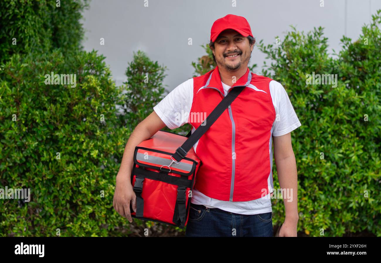 Un uomo sorridente addetto alla consegna in maglietta con cappuccio rosso, borsa termica per alimenti, zaino, servizio di corriere in piedi sullo sfondo della natura Foto Stock