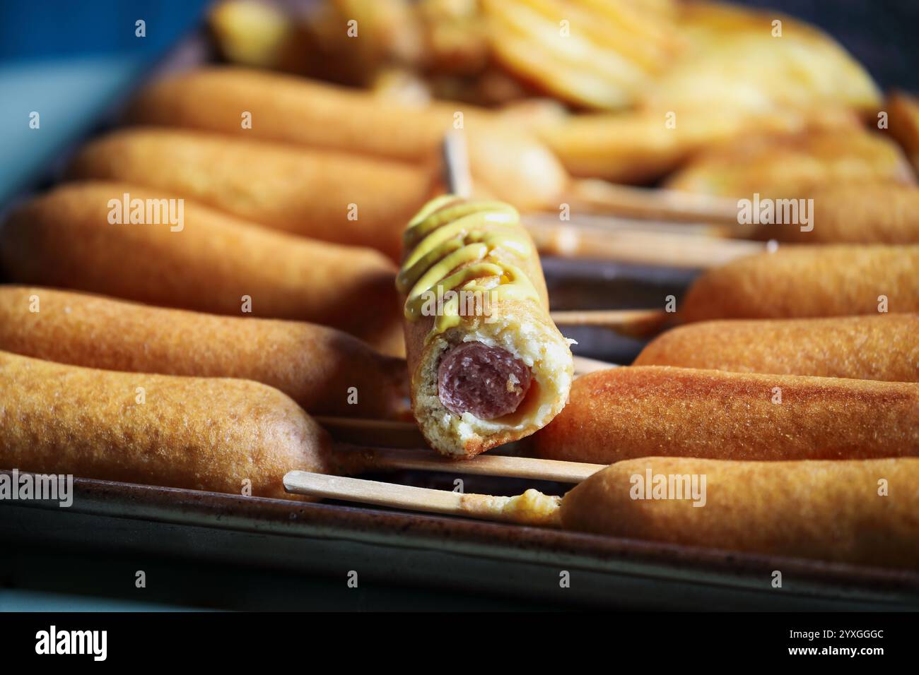Padella piena di cani da mais, uno con senape e patatine fritte sullo sfondo. Messa a fuoco selettiva con sfondo sfocato. Foto Stock