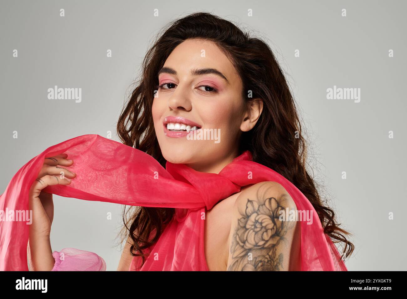 Una giovane donna con i capelli lunghi e un tatuaggio floreale sorride, tenendo elegantemente una sciarpa rosa. Foto Stock