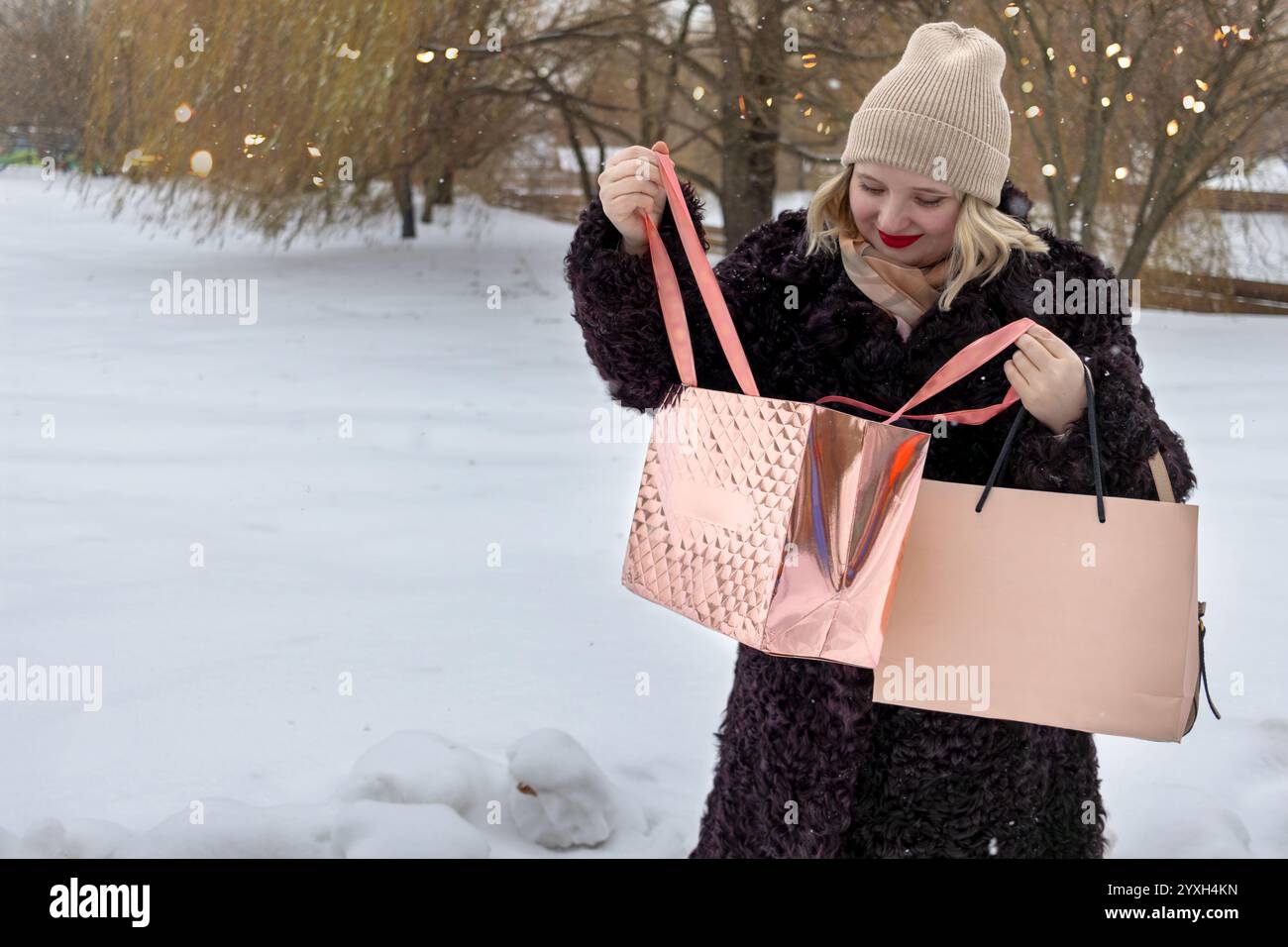 Una donna in pelliccia e cappello sbircia in borse piene di regali di Natale mentre cammina lungo le strade innevate invernali. Shopping di Natale. Spazio di copia, per il testo. Foto Stock