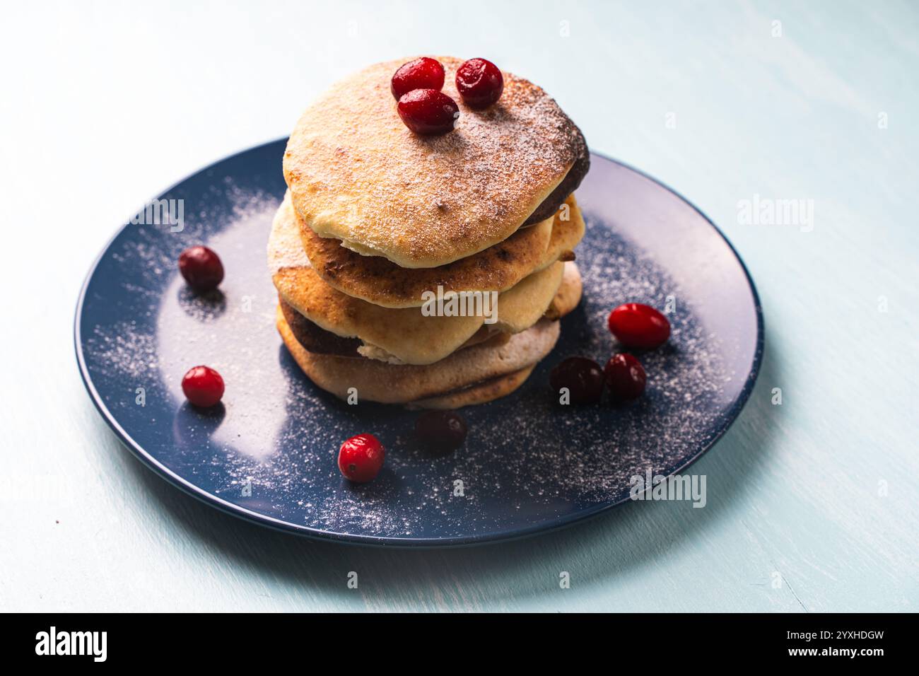 Le frittelle di formaggio con mirtilli rossi sono impilate su un piatto blu su un tavolo azzurro, cosparse di zucchero a velo. Foto di alta qualità Foto Stock
