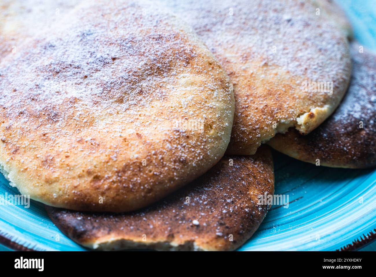 Le frittelle di formaggio con mirtilli rossi sono impilate su un piatto blu su un tavolo azzurro, cosparse di zucchero a velo. Foto di alta qualità Foto Stock