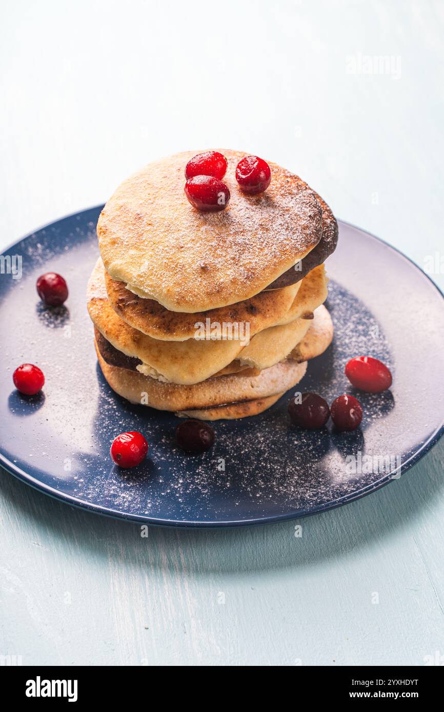 Le frittelle di formaggio con mirtilli rossi sono impilate su un piatto blu su un tavolo azzurro, cosparse di zucchero a velo. Foto di alta qualità Foto Stock