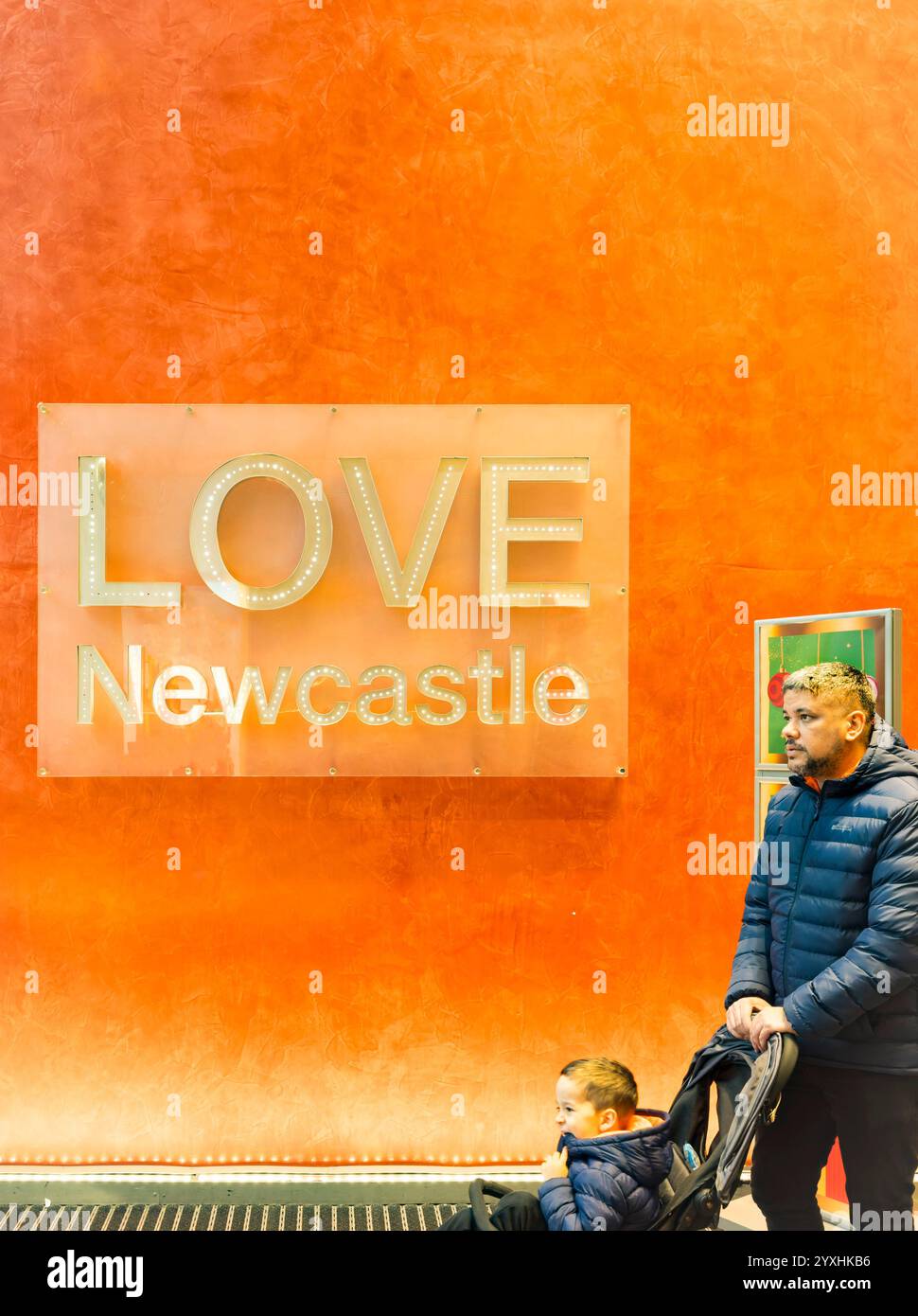 Uomo che spinge un passeggino con un bambino davanti a un muro arancione con un cartello "LOVE Newcastle", che mette in risalto l'orgoglio locale e la vita urbana Foto Stock
