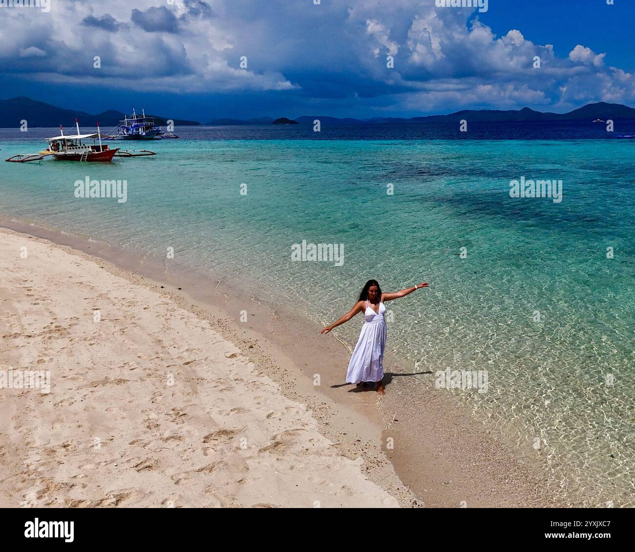 Una splendida giovane donna in un abito bianco che scorre si erge graziosamente su sabbia bianca, con acqua turchese e barche ormeggiate sullo sfondo, evocando un se Foto Stock