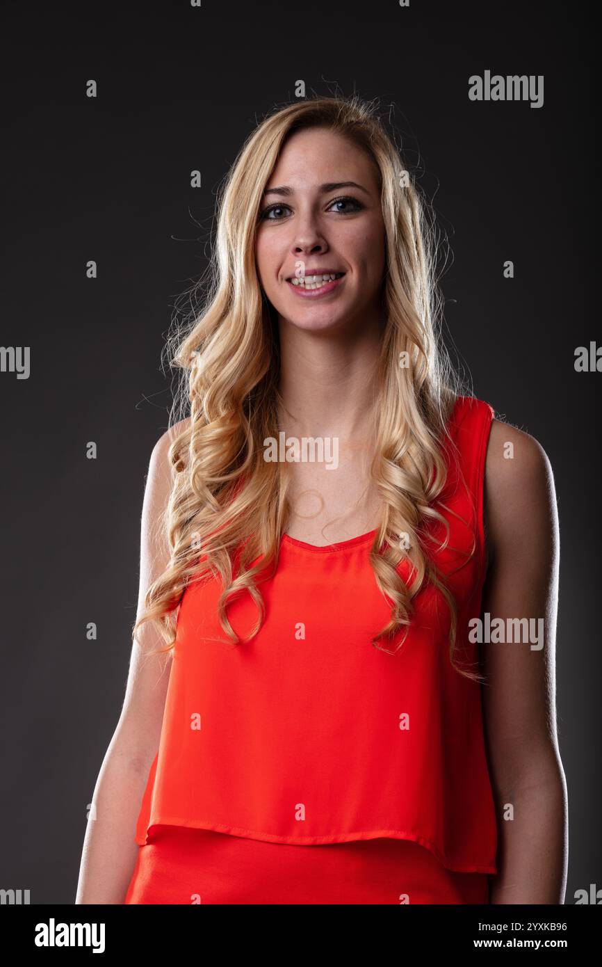 Una donna bionda con un vestito arancione, catturata in un ritratto a mezza lunghezza. Sorriso delicato, braccia rilassate, sfondo nero, accentuato dalla luce da destra Foto Stock