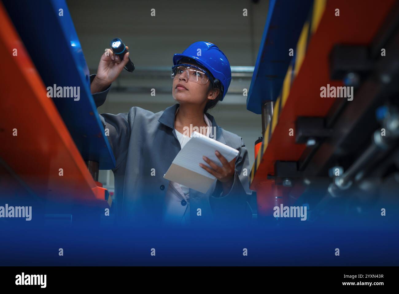 Ingegnere donna indiana che lavora in una linea di produzione, esaminando il lavoro di macchinari automatizzati e scrivendo una lista di controllo per la pianificazione. Foto Stock