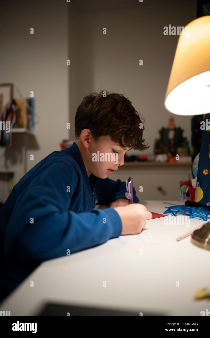 Ragazzo di 11 anni seduto alla sua scrivania in camera da letto scrivendo biglietti d'auguri di Natale, Inghilterra, Regno Unito Foto Stock