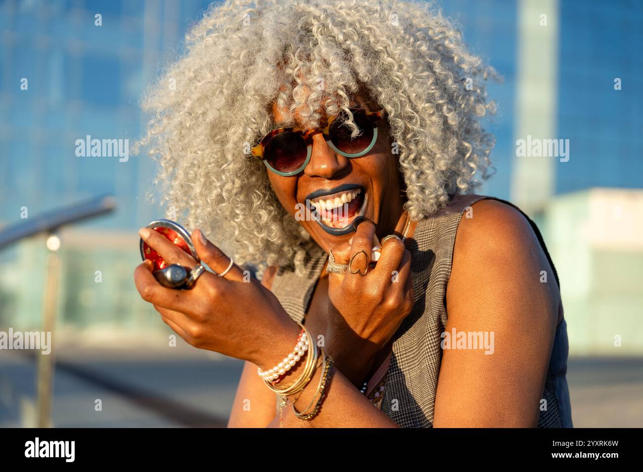 Signora nera espressiva con capelli afro e occhiali da sole che indossano rossetto blu nel soleggiato centro della città. Concetto: pro-aging, fiducia in se stessi Foto Stock