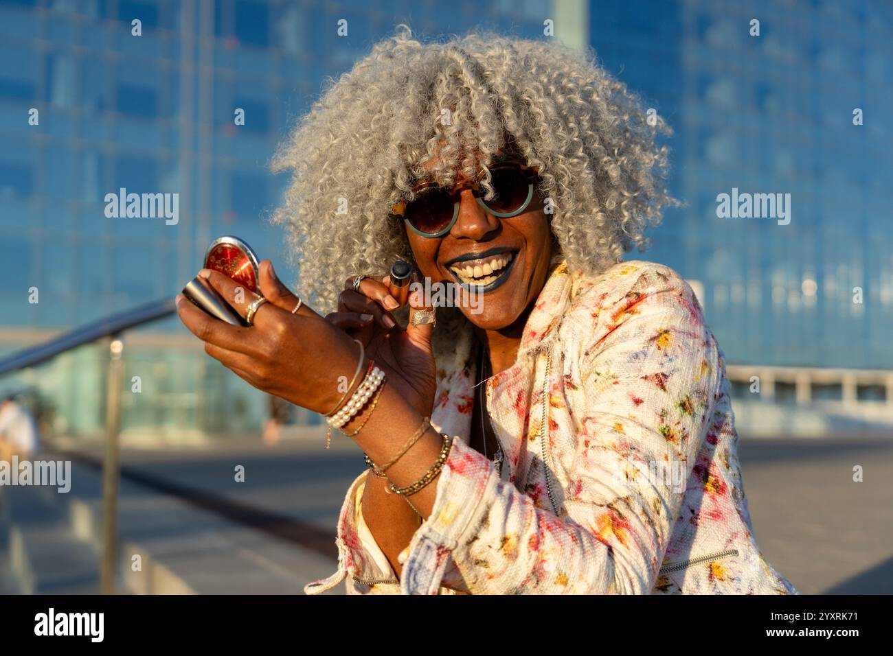Signora nera espressiva con capelli afro e occhiali da sole che indossano rossetto blu nel soleggiato centro della città. Concetto: pro-aging, fiducia in se stessi Foto Stock