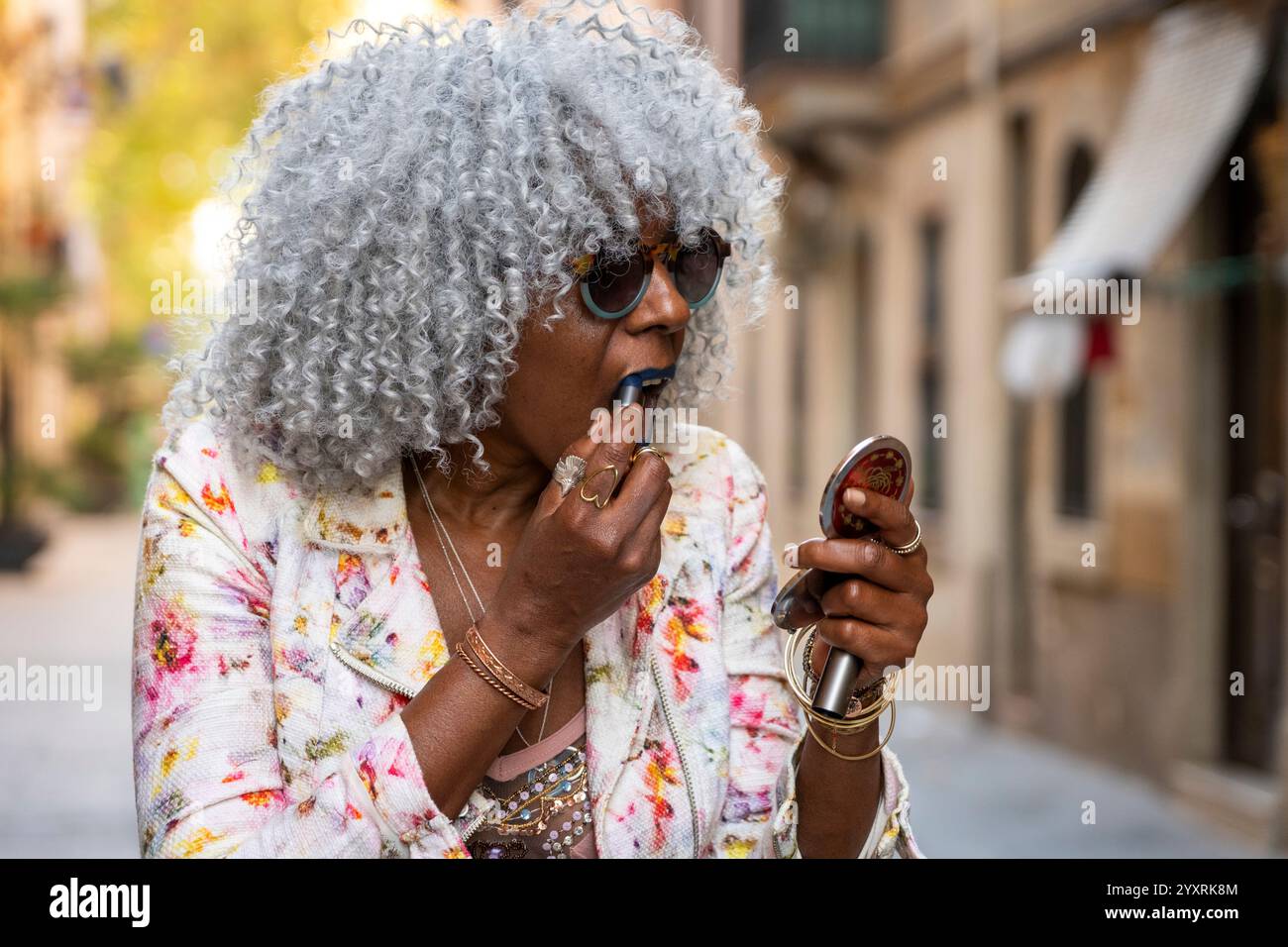 Signora nera espressiva con capelli afro e occhiali da sole che indossano rossetto blu nel soleggiato centro della città. Concetto: pro-aging, fiducia in se stessi Foto Stock
