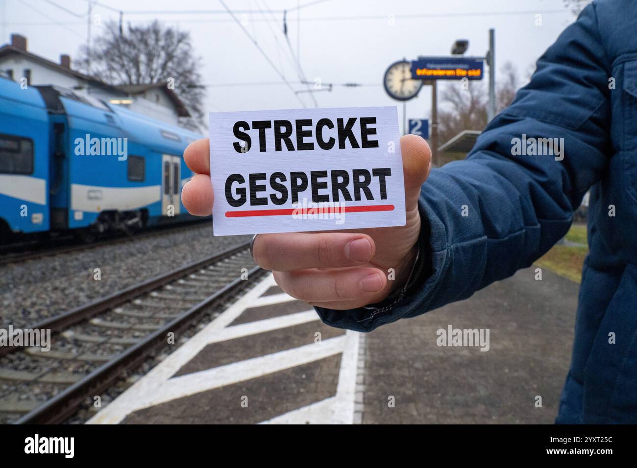 Baviera, Germania - 17 dicembre 2024: Una persona ha un cartello con il testo STRECKE GESPERRT davanti a una stazione e ai binari. Immagine simbolica per chiusure di linee, lavori di costruzione o incidenti nel traffico ferroviario. FOTOMONTAGGIO *** Eine person hält ein Schild mit dem Text STRECKE GESPERRT vor einem Bahnhof und Gleisen. Symbolbild für Streckensperrungen, Bauarbeiten oder Unfälle im Schienenverkehr. FOTOMONTAGE Foto Stock