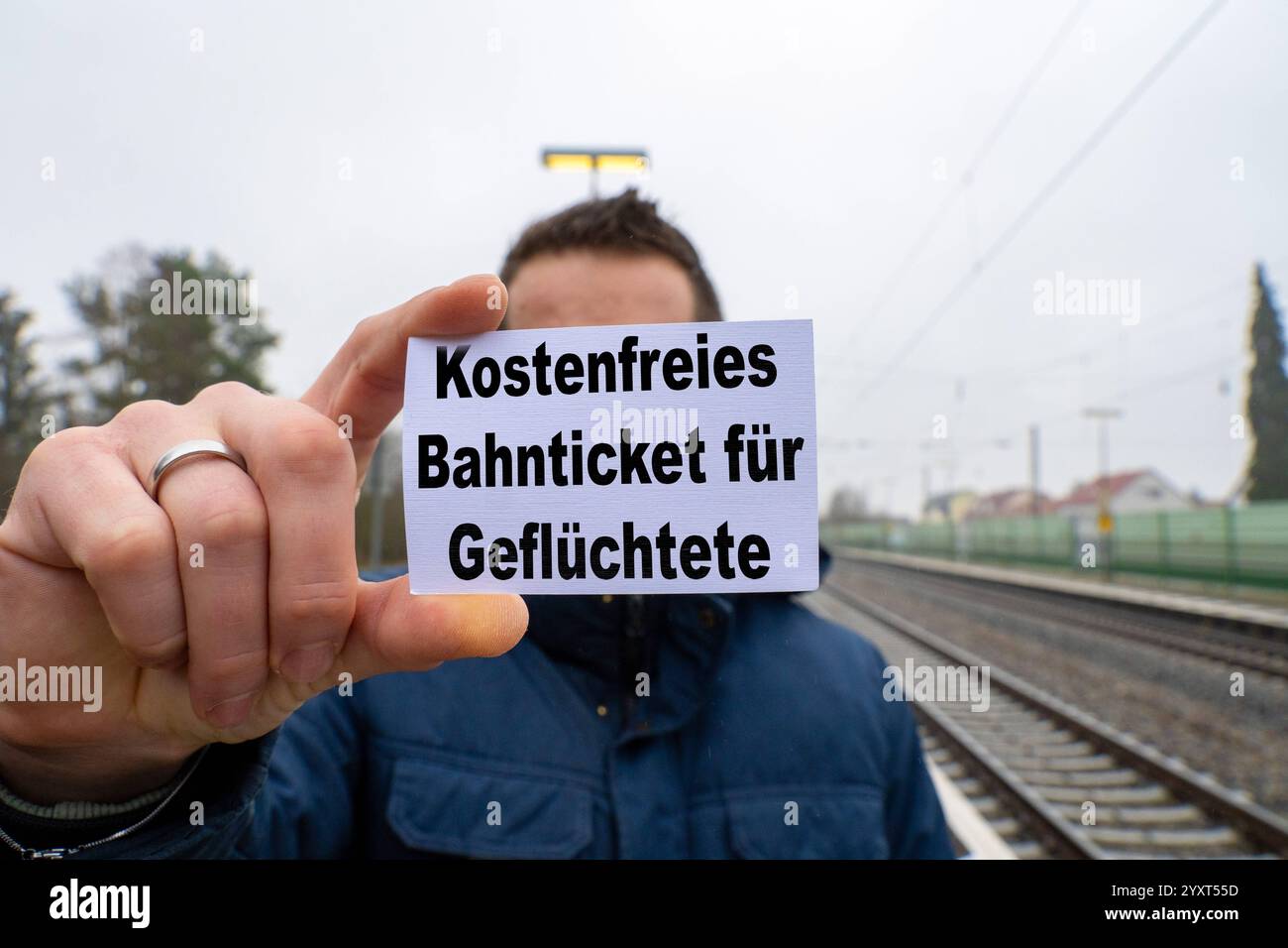 Baviera, Germania - 17 dicembre 2024: Una persona in possesso di un cartello con il BIGLIETTO DEL TRENO GRATUITO PER I RIFUGIATI DI fronte a una stazione ferroviaria e ai binari. Immagine simbolica delle misure di aiuto umanitario, del sostegno ai rifugiati ucraini e della libera mobilità per le persone che cercano protezione. FOTOMONTAGGIO *** Eine person hält ein Schild mit dem Text KOSTENFREIES BAHNTICKET für GEFLÜCHTETE vor einem Bahnhof und Gleisen. Symbolbild für humanitäre Hilfsmaßnahmen, Unterstützung für Geflüchtete aus der Ukraine und kostenlose Mobilität für Schutzsuchende. FOTOMONTAGE Foto Stock