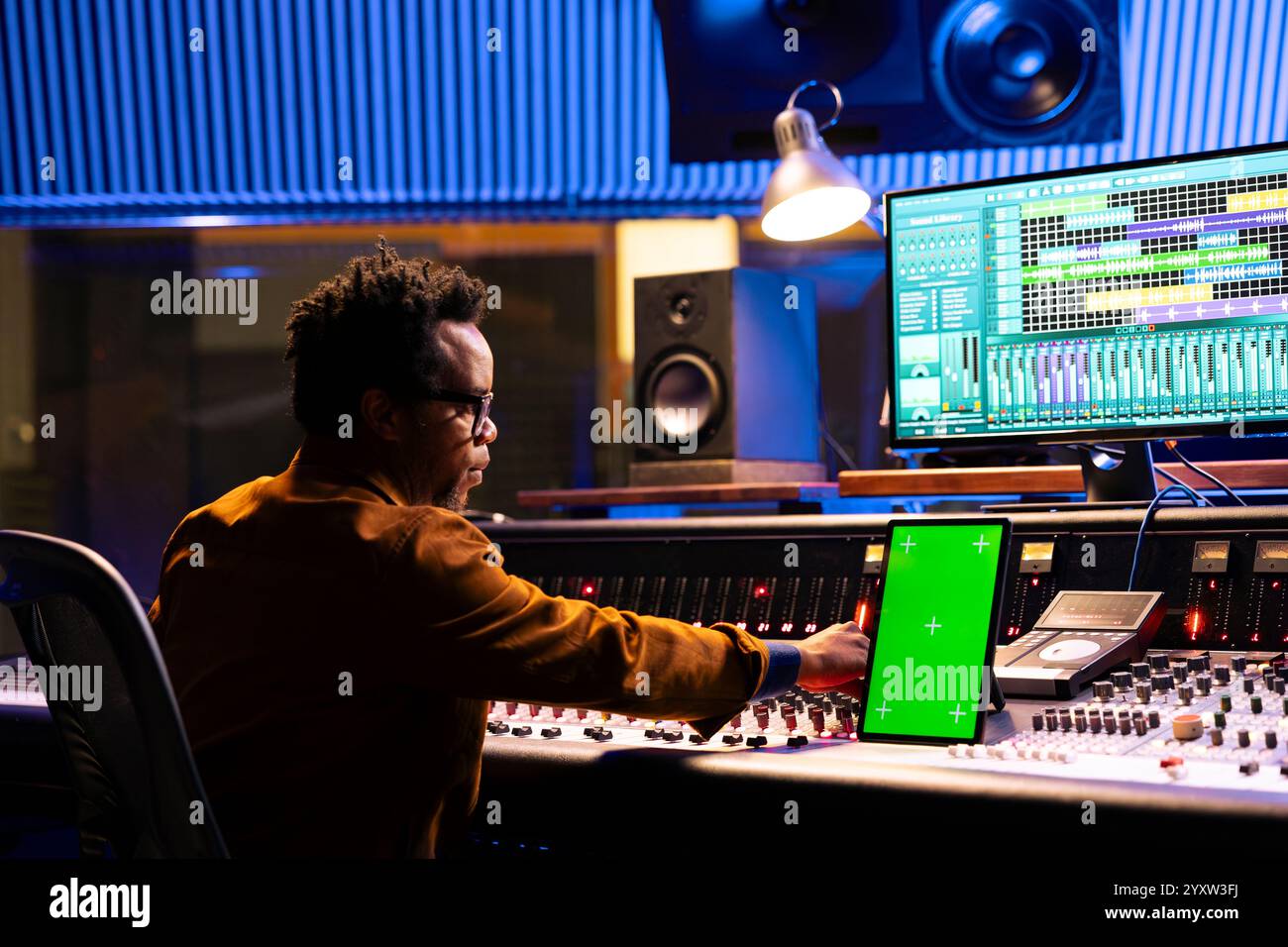 I tecnici del suono mescolano le tracce accanto al tablet con schermo verde nella sala di controllo, le manopole e i fader della scheda del quadro di comando. Produttore afroamericano che registra canzoni di successo ed elabora suoni. Foto Stock