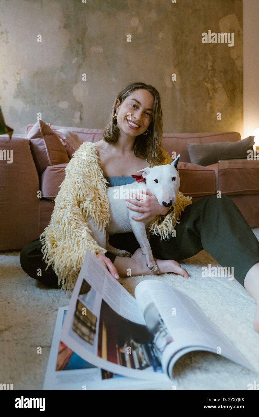 Ritratto di una giovane donna sorridente seduta sul pavimento con un cane e che tiene una rivista vicino al divano di casa Foto Stock