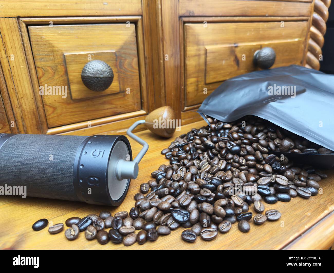 Primo piano di chicchi di caffè freschi interi che fuoriescono da una confezione grande aperta, accanto a un macinacaffè manuale su un armadietto di pini. Foto Stock