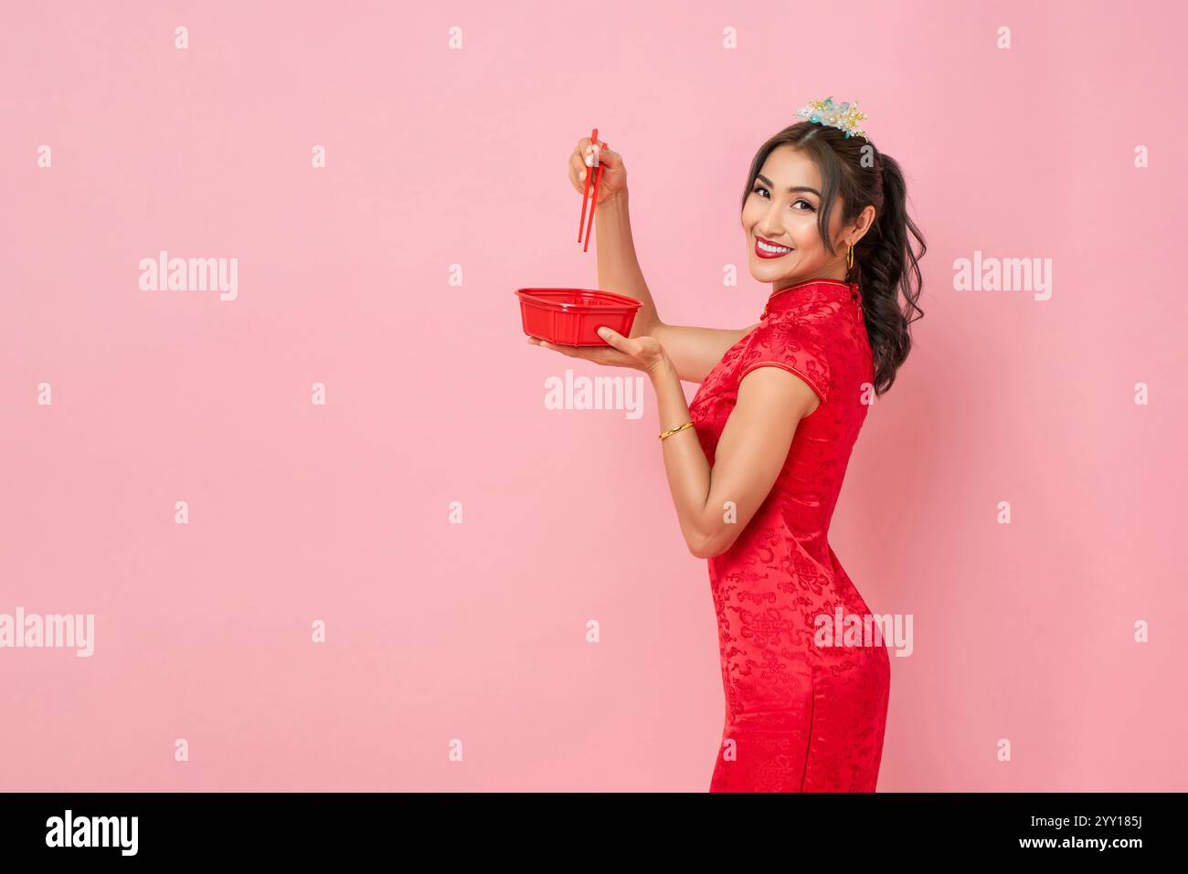 Donna asiatica in tradizionale abito qipao rosso cinese con scatola di plastica e bacchette su sfondo rosa isolato in studio Foto Stock